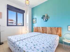 Quarto com duas camas, cabeceira de madeira, janela com vista para edifícios e parede azul-turquesa com quadros.
