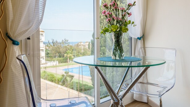 Un balcón con una mesa de vidrio, un jarrón con flores y vistas a una piscina y vegetación.