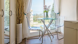 Un balcón con una mesa y sillas de vidrio, un florero con flores y vista al mar.