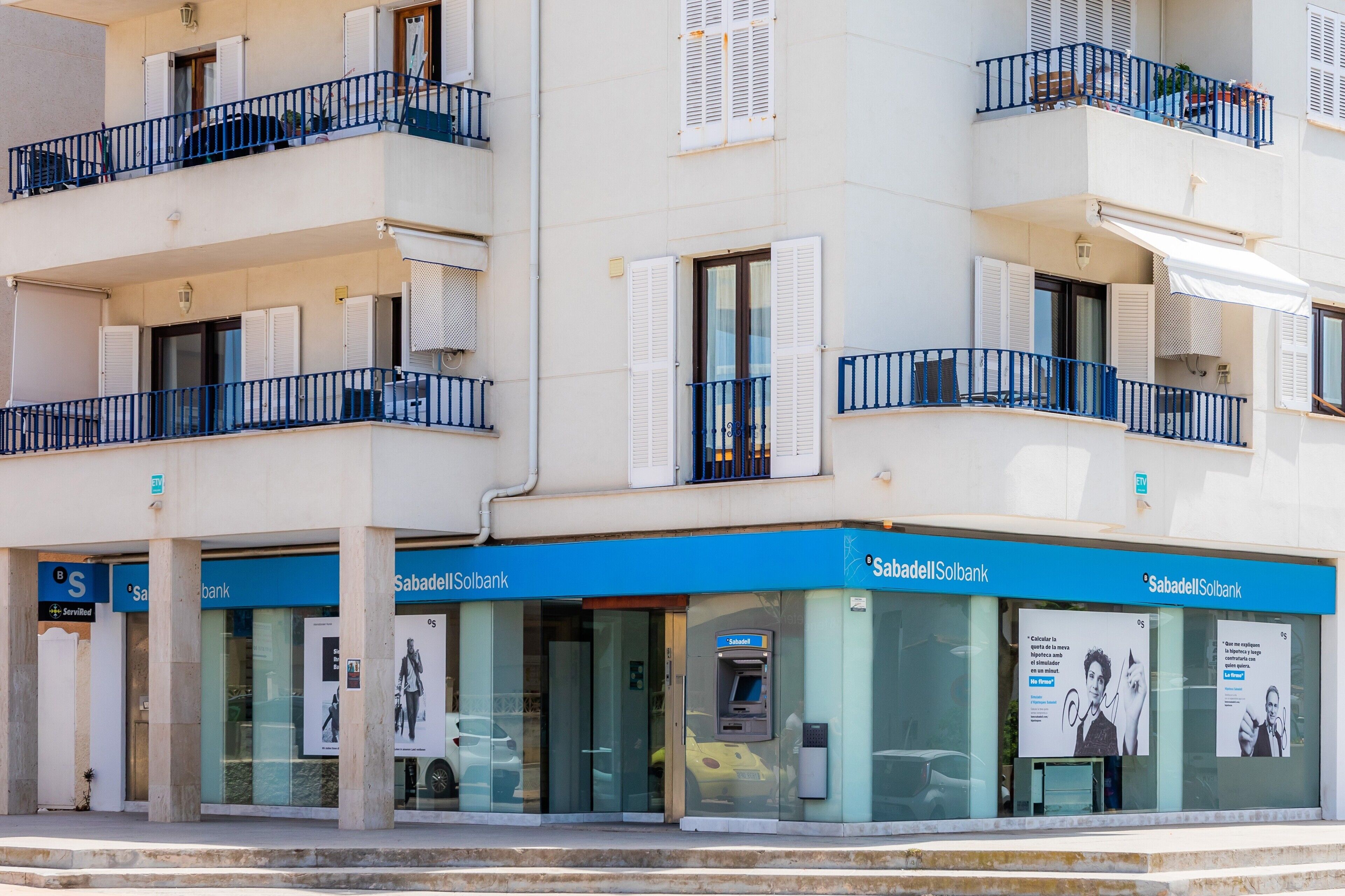 En bygning med flere etager, hvor der i stueetagen er en bank, SabadellSolbank, med glasdøre, en pengeautomat og en blå markise.
