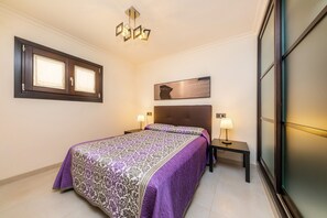 Une chambre à coucher comprenant un lit, deux lampes de chevet, une table de chevet, une fenêtre avec des stores, un tableau au mur et un luminaire au plafond.
