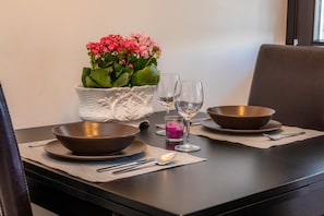 Een eettafel gedekt met twee kommen, een glas paars drankje en een witte vaas met roze bloemen.