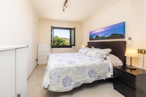 A bedroom with a bed, a nightstand, a lamp, a window with a view, and a painting on the wall.