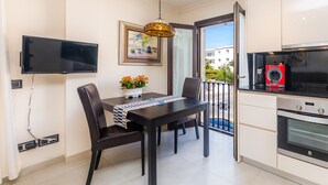 Un espace repas comprenant une table, des chaises, un téléviseur fixé au mur, et une cuisine équipée d’un four et d’une machine à café.