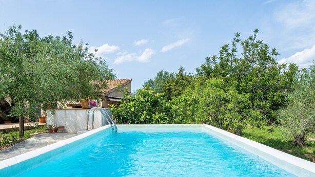 Una piscina con agua cristalina azul, rodeada de vegetación y un edificio al fondo.