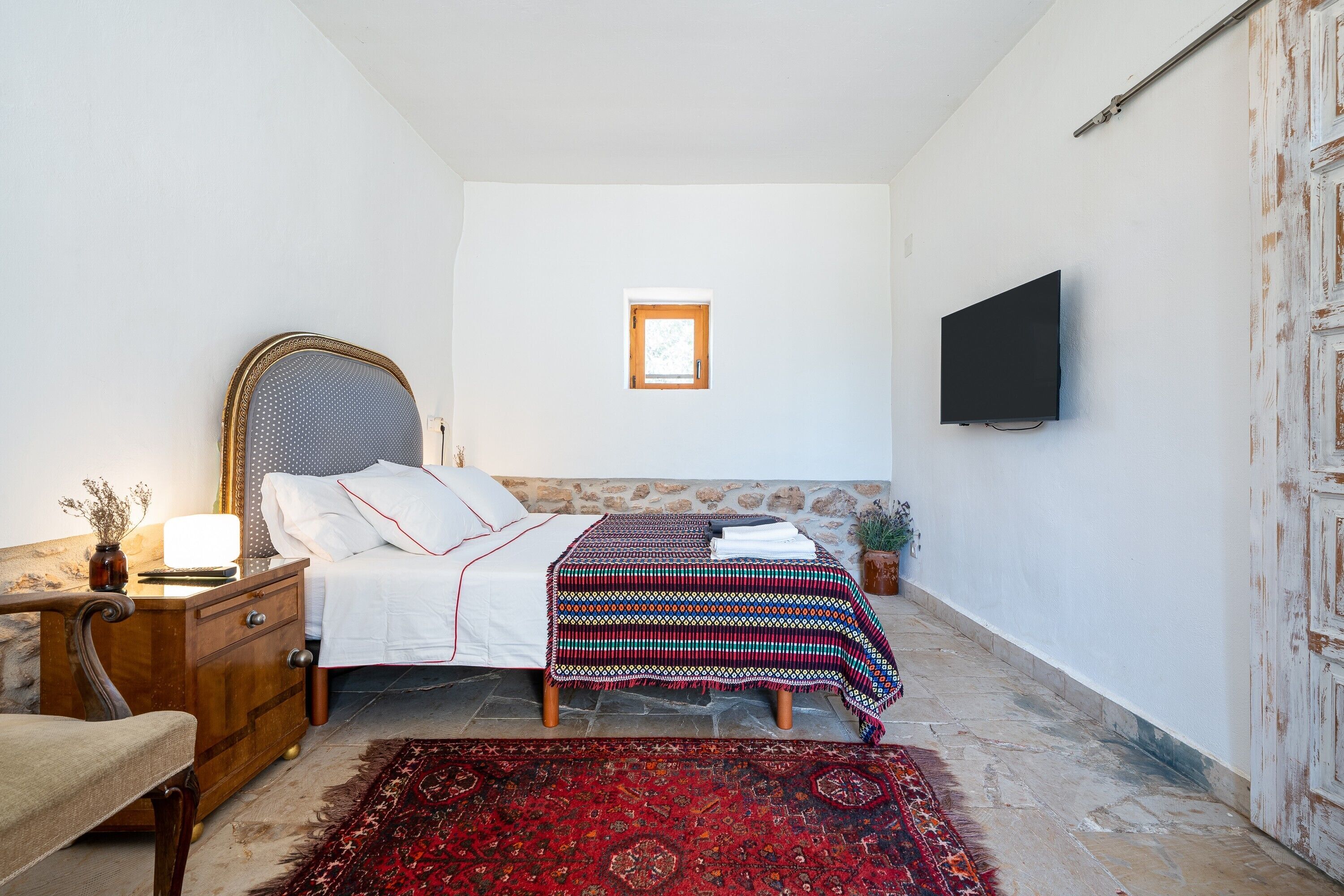 Un dormitorio con cama, mesita de noche, lámpara, televisor y un ventanal.