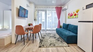 Ein modernes Wohnzimmer mit einem blauen Sofa, einem Essbereich mit orangefarbenen Stühlen, einem an der Wand befestigten Fernseher und einem großen Fenster mit transparenten Vorhängen.