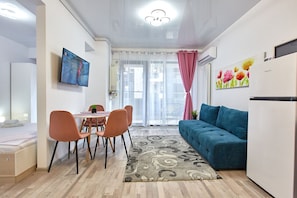 A modern living room with a blue sofa, a dining area with orange chairs, a TV mounted on the wall, and a large window with sheer curtains.