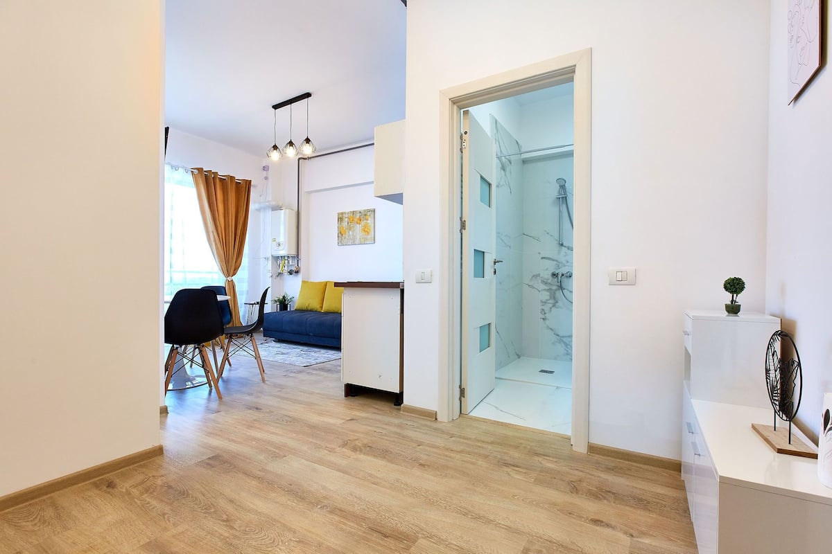 Un salon moderne avec un canapé, une table à manger et des chaises. On aperçoit une porte de douche en verre à l’arrière-plan.