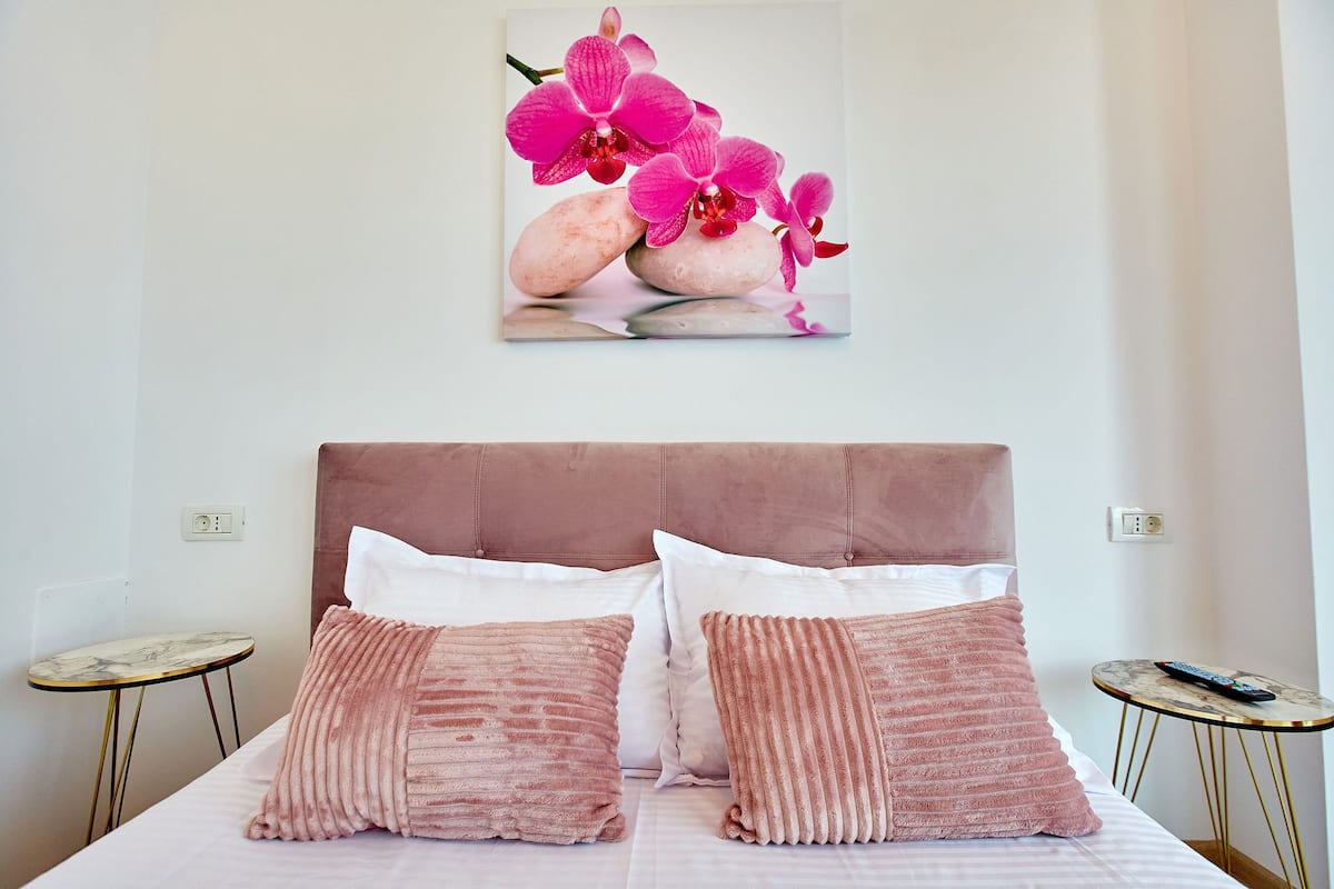 Une chambre avec un grand lit, deux tables de chevet et une œuvre d’art représentant des orchidées roses, accrochée au mur.