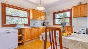 A kitchen with wooden cabinets, a washing machine, a dining table with a fruit plate, and a coffee maker.