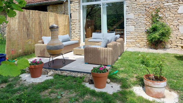 Een stenen huis met een pannendak, een grote glazen deur en een terras met rieten meubels en potplanten.