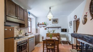 Een keuken met houten kastjes, een eettafel met stoelen, een hanglamp en een aan de muur bevestigde televisie.