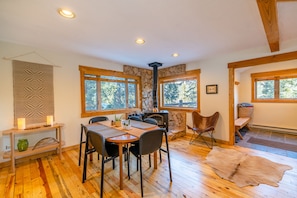 A cozy room with a stone wall, wooden beams, a fireplace, a dining table with chairs, and large windows.