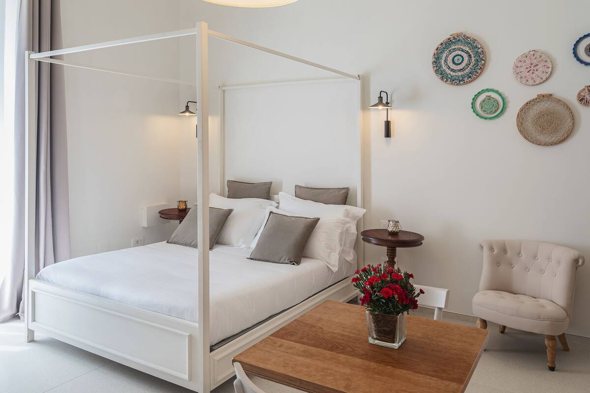 A four-poster bed with a wooden headboard, a small round table with a vase of red flowers, a white armchair, and decorative wall hangings.
