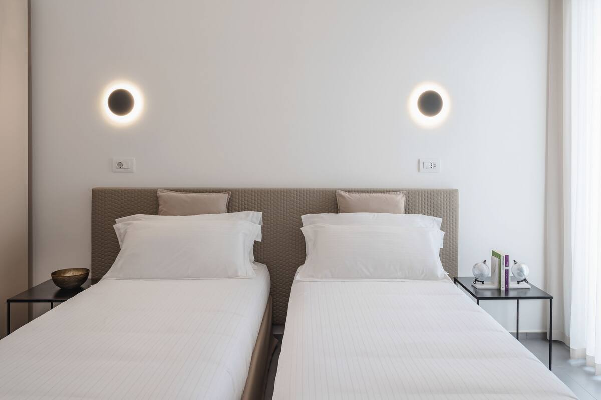 A hotel room with two beds, a small table with a bowl and books, and two wall-mounted lights.