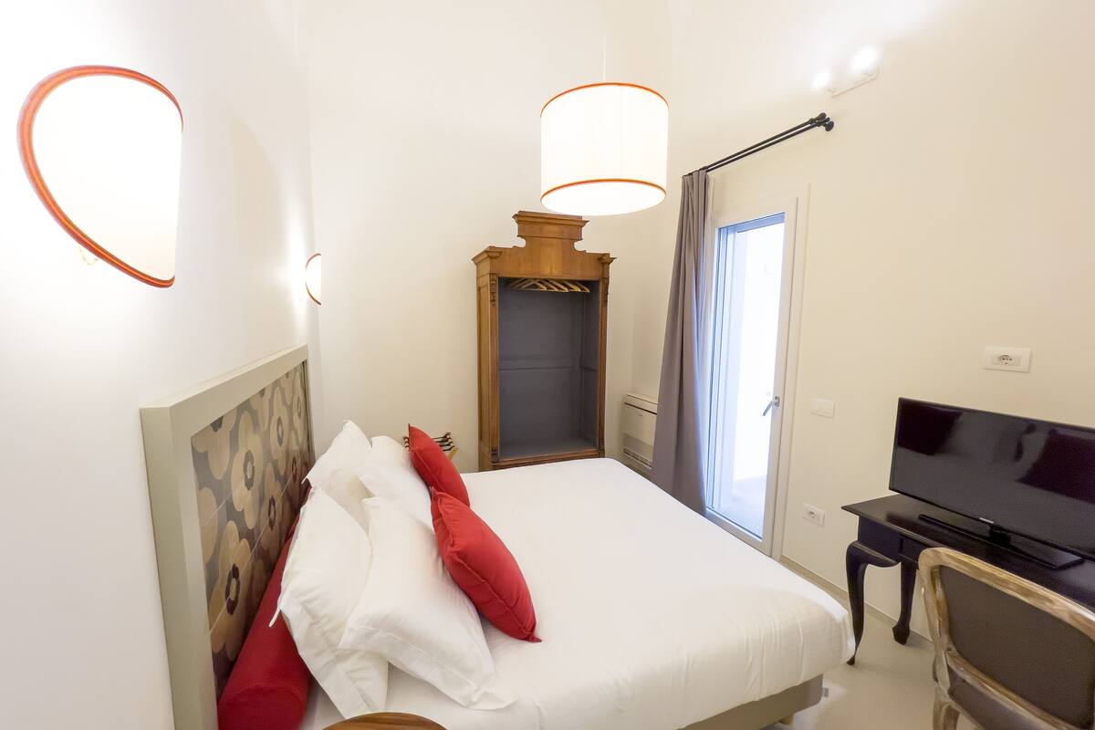 A hotel room with a bed, a desk with a television, a wardrobe, and a window with curtains.