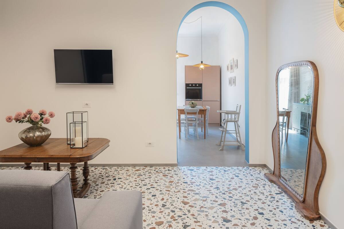 A modern living room with a wooden dining table, a sofa, a TV mounted on the wall, and a large mirror.