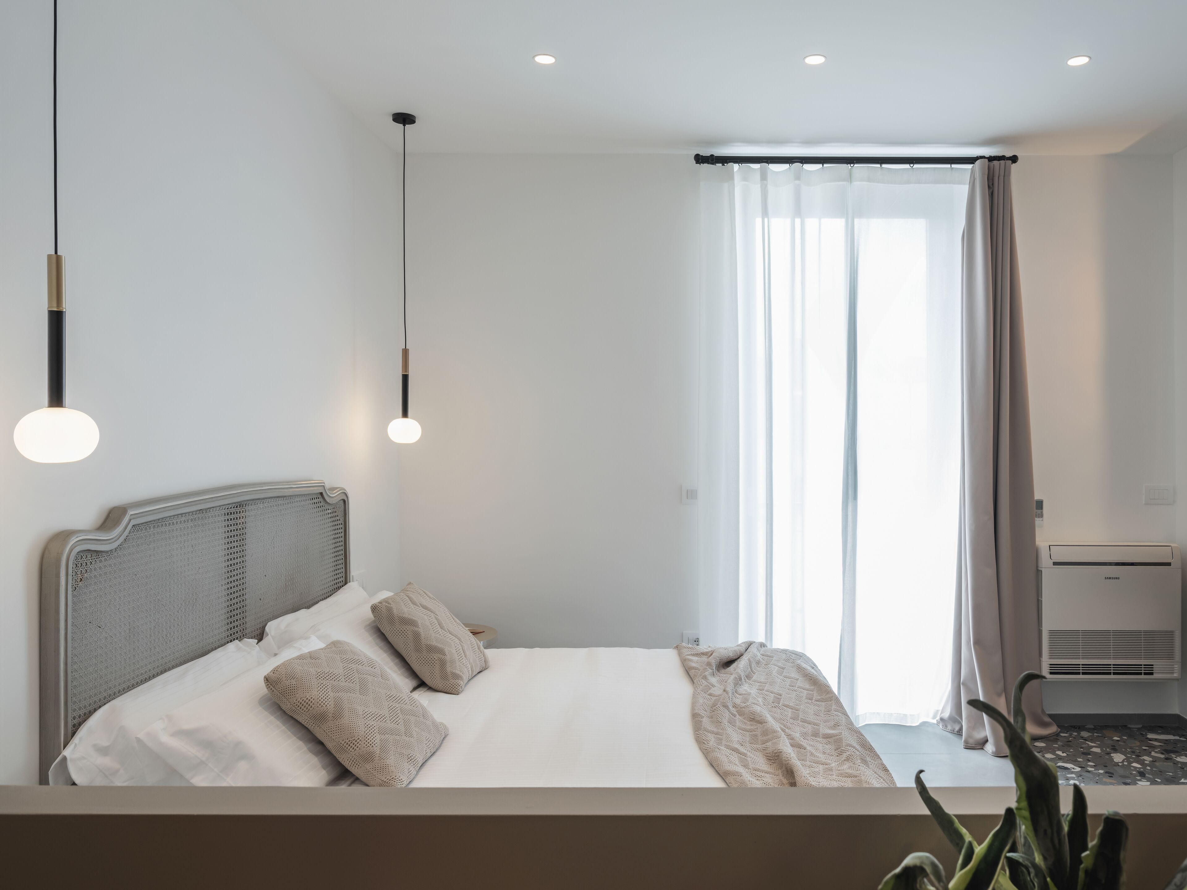 A modern hotel room with a large bed, a bedside table, a wall-mounted air conditioner, and a window with sheer curtains.