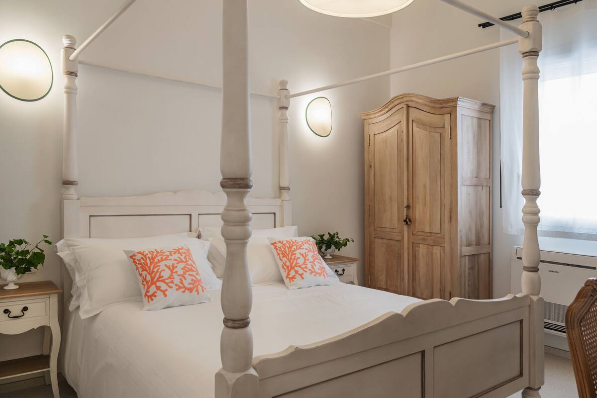 A four-poster bed with white bedding and orange decorative pillows, a wooden wardrobe, and a bedside table with a plant.