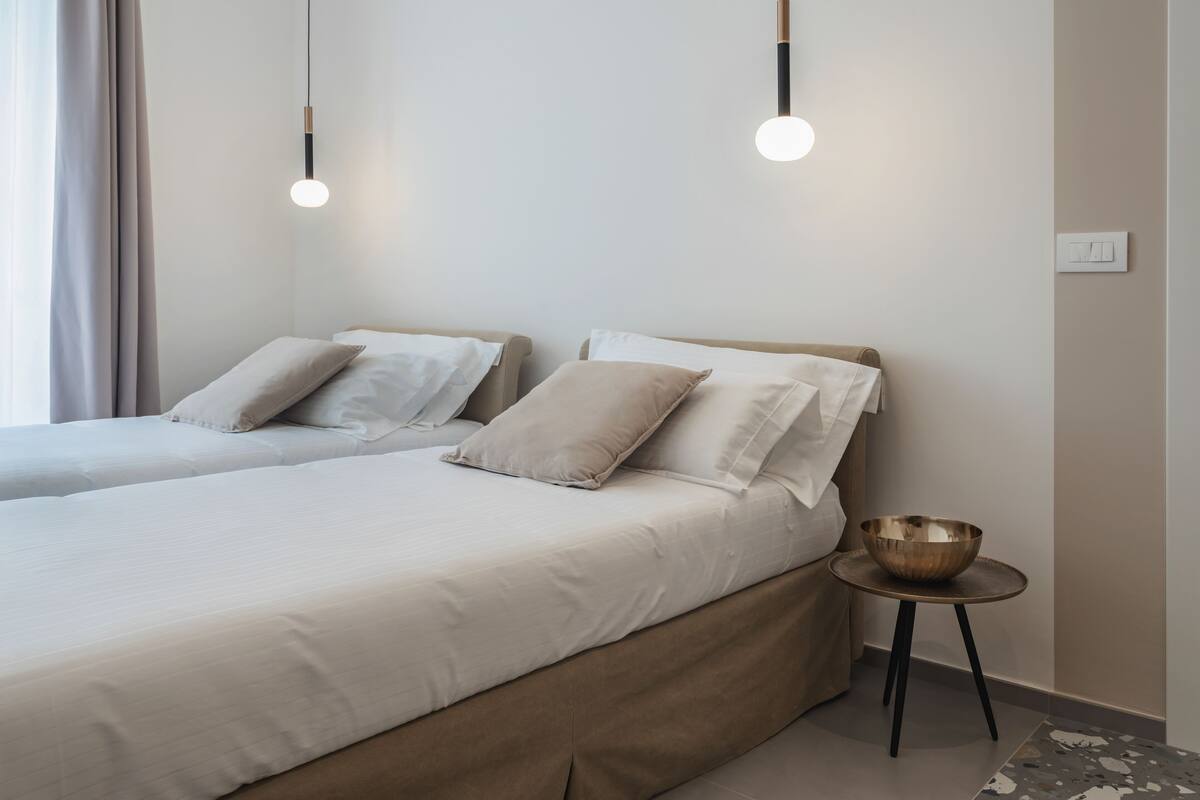 A modern bedroom with two beds, a bedside table with a bowl, and pendant lights.