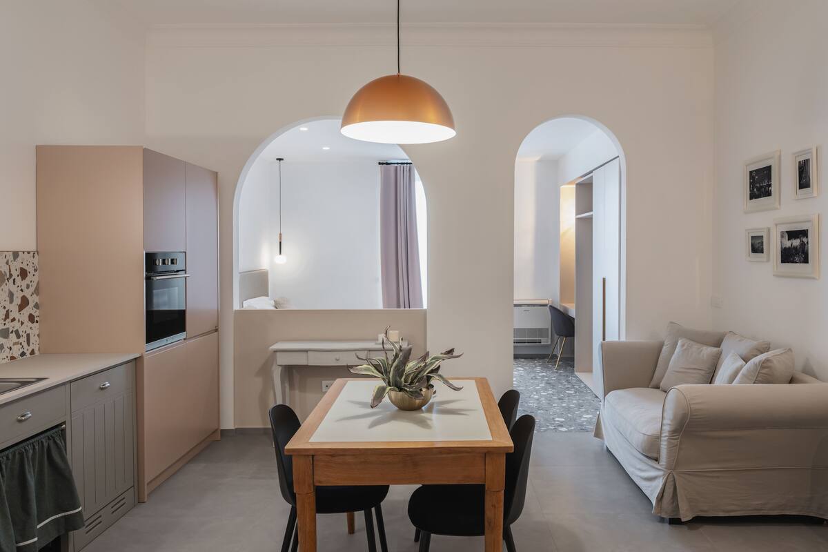 A modern kitchen with an oven, a dining table with chairs, and a pendant light. There is a sofa and a coffee table in the living area.