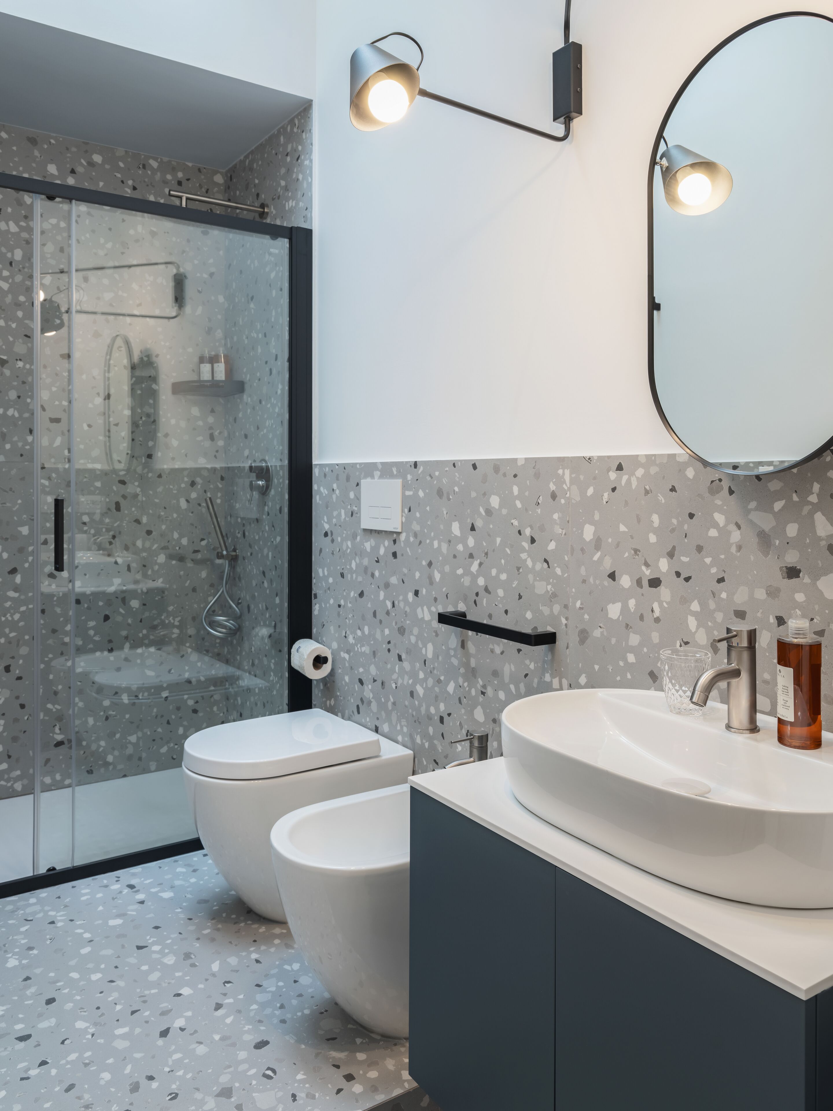 Un baño moderno con cabina de ducha de vidrio, inodoro blanco, lavamanos grande con espejo y suelo de terrazo.