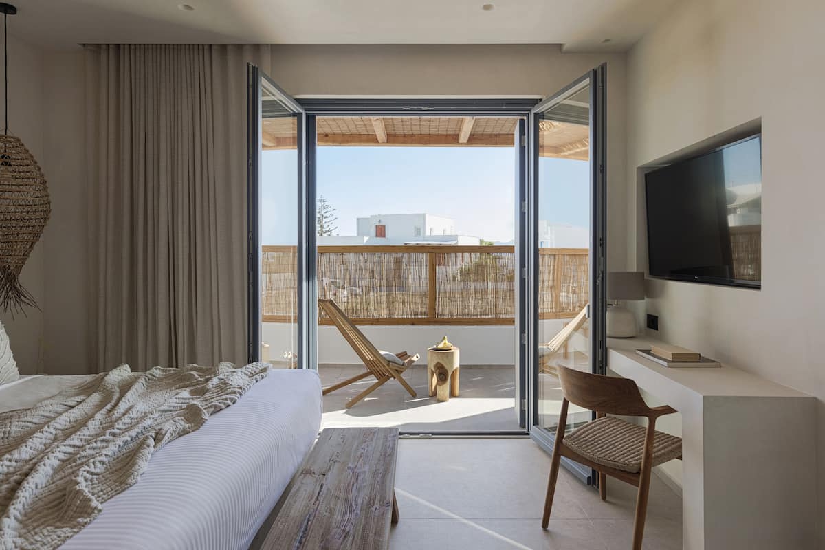 A modern bedroom with a large bed, a wooden desk, a TV, and a balcony with a chair and a small table.