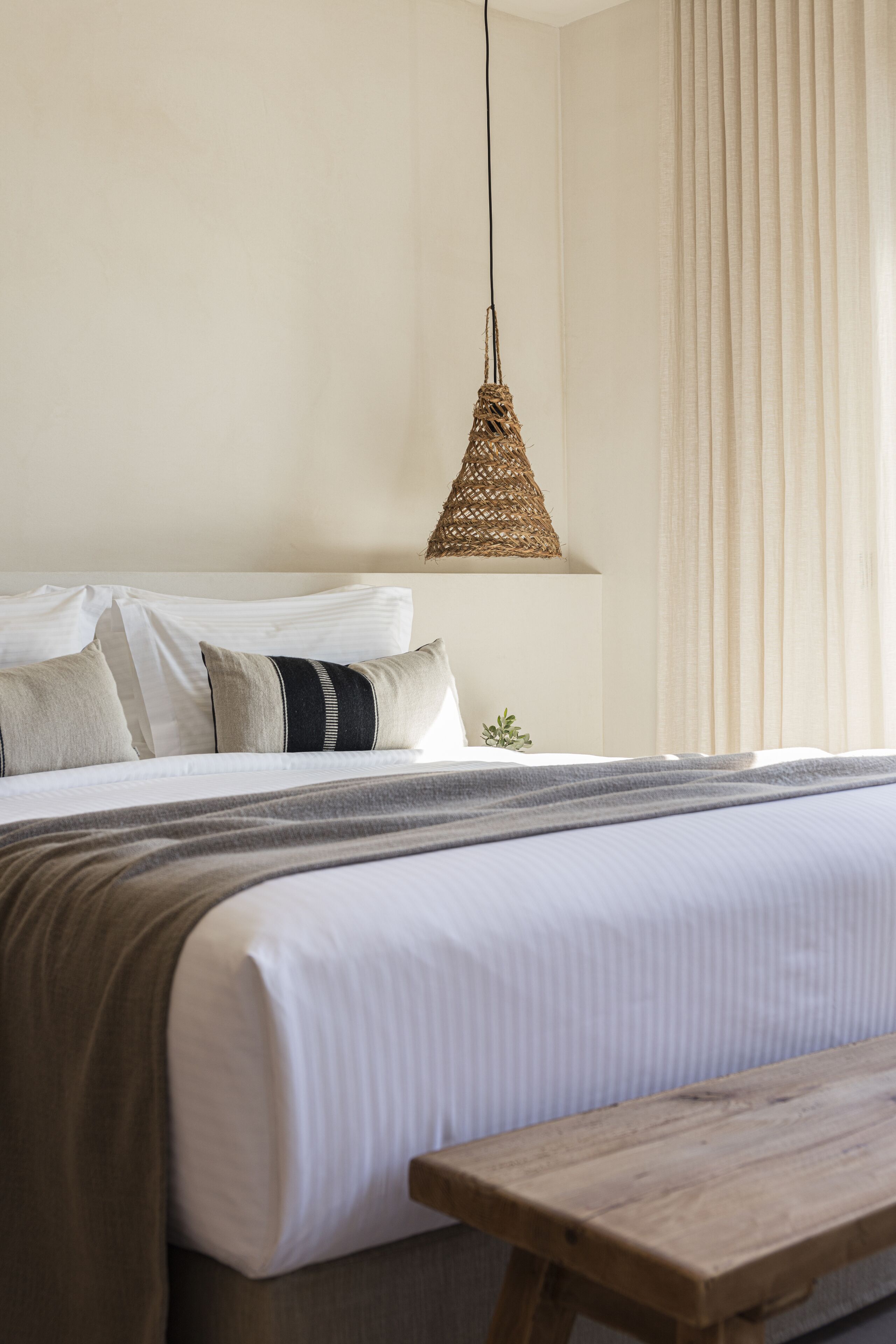Una cama bien tendida con un cabecero de madera, una lámpara colgante tejida y vistas a una ventana con cortinas.