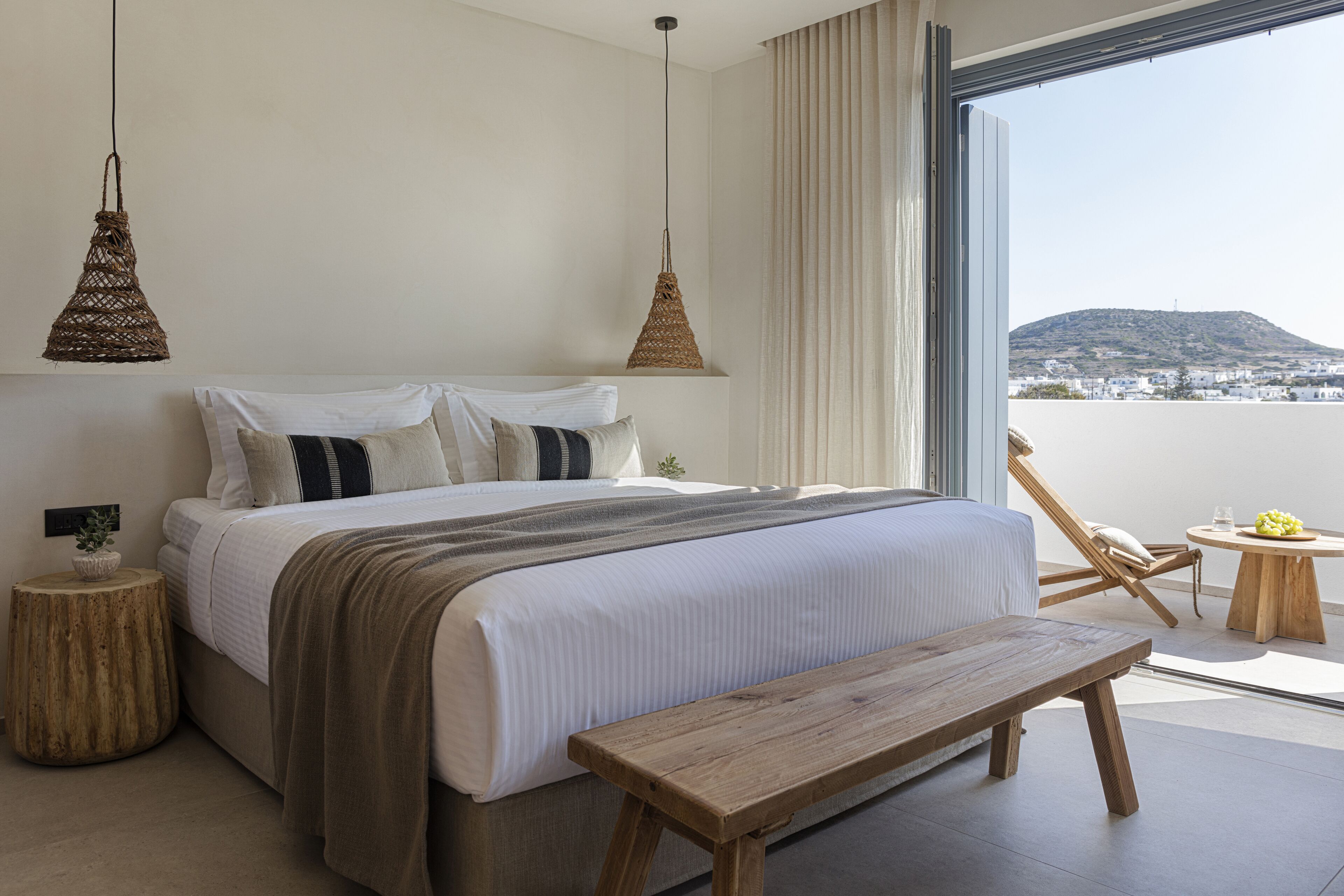 A modern bedroom with a large bed, two hanging lamps, a wooden bench, and a view of a distant landscape through a large window.