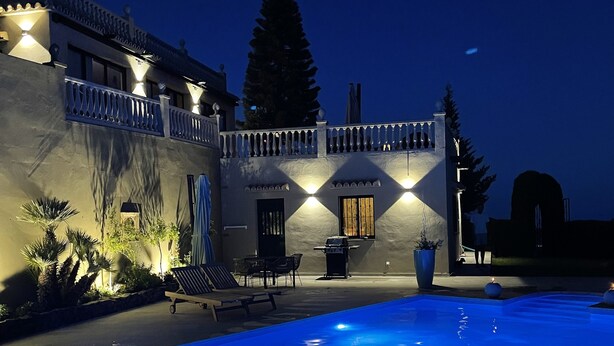Zona d'una piscina de nit amb un edifici que té un balcó i zones d'estar a l'aire lliure.