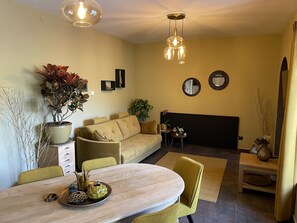 Een moderne woonkamer met een bank, eettafel en stoelen. Er staan plantenpotten, hangt kunst aan de muur en staat een televisie.