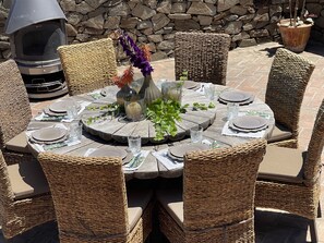 Een ronde eettafel buiten, compleet met rieten stoelen, een stenen muur op de achtergrond en een barbecue.