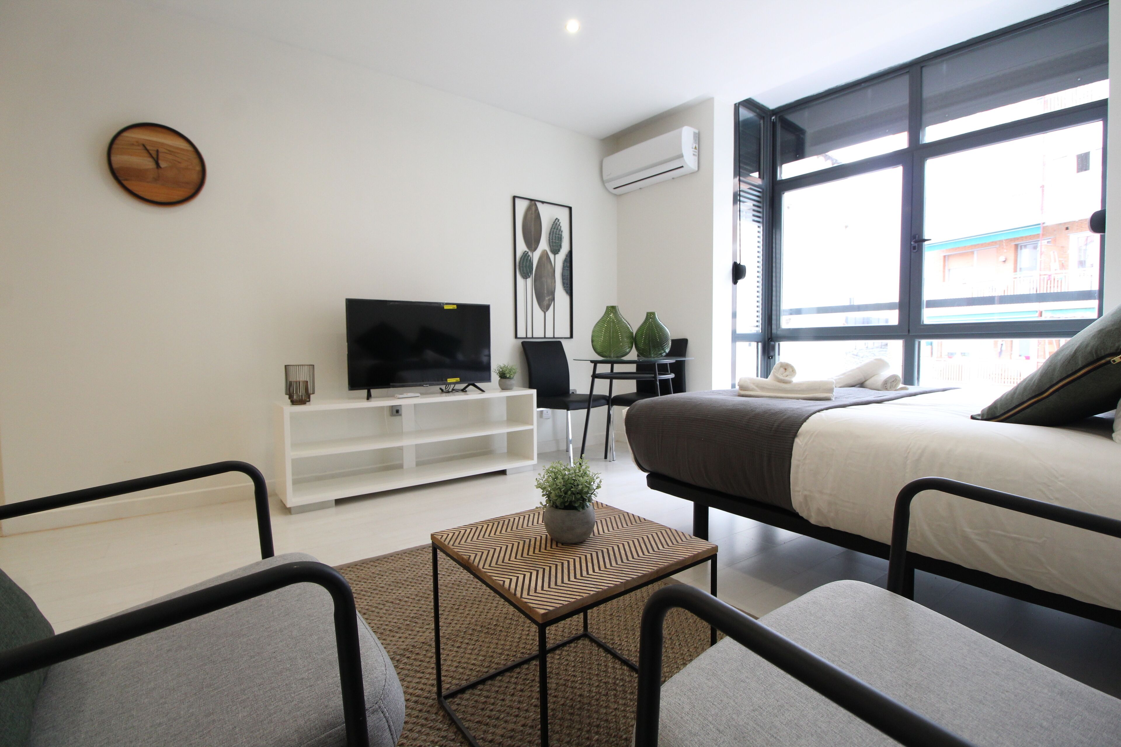 A modern hotel room with a bed, a TV, a small table with a plant, and a window with a view of buildings.