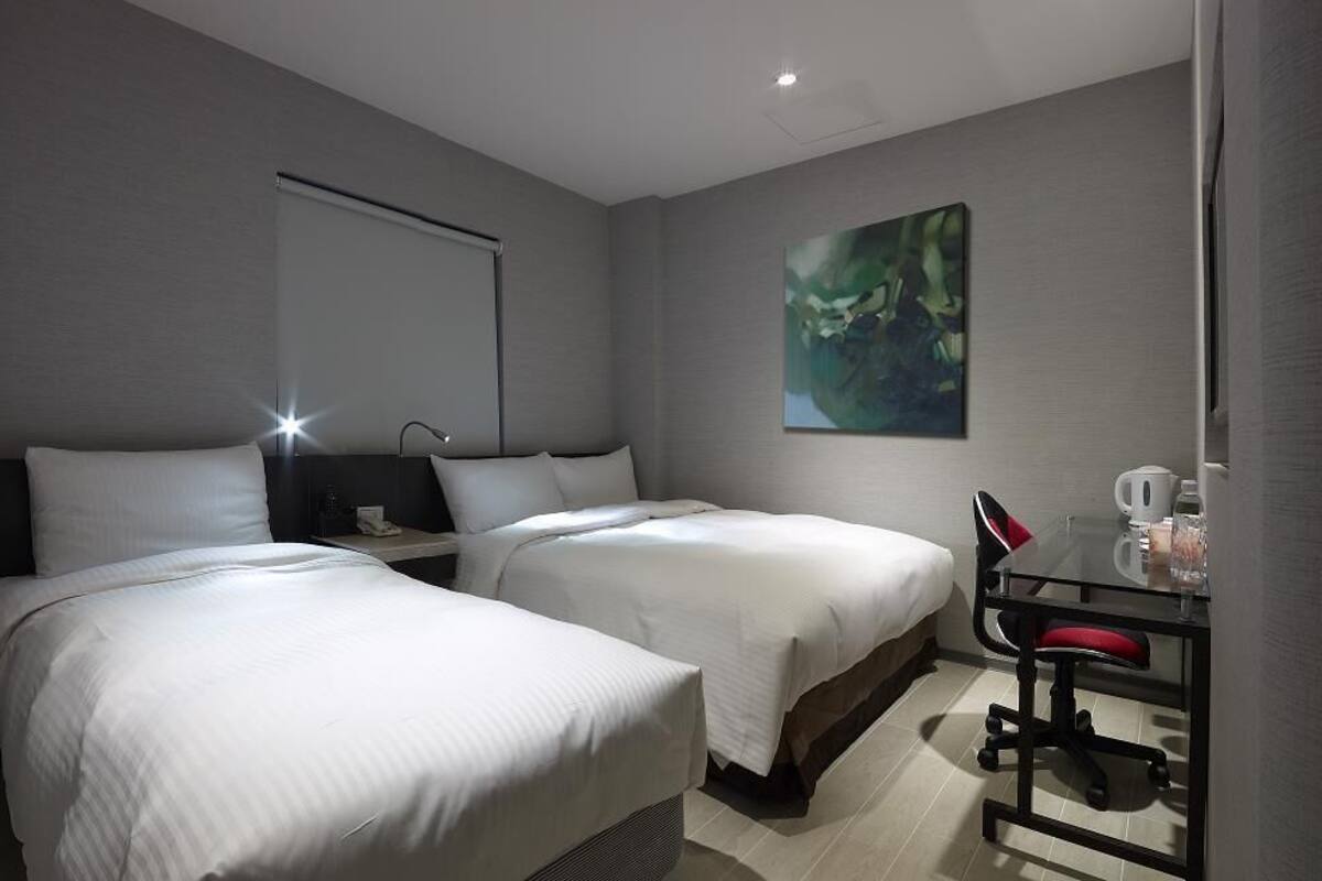 A modern hotel room with two single beds, a desk with a chair, a coffee maker, and a painting on the wall.