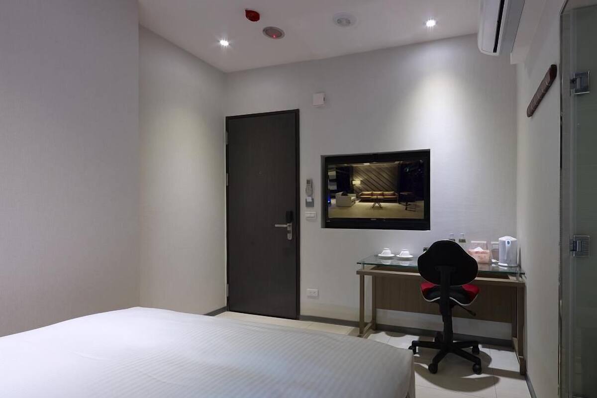 A hotel room with a bed, a desk with a chair, a television mounted on the wall, and a door.