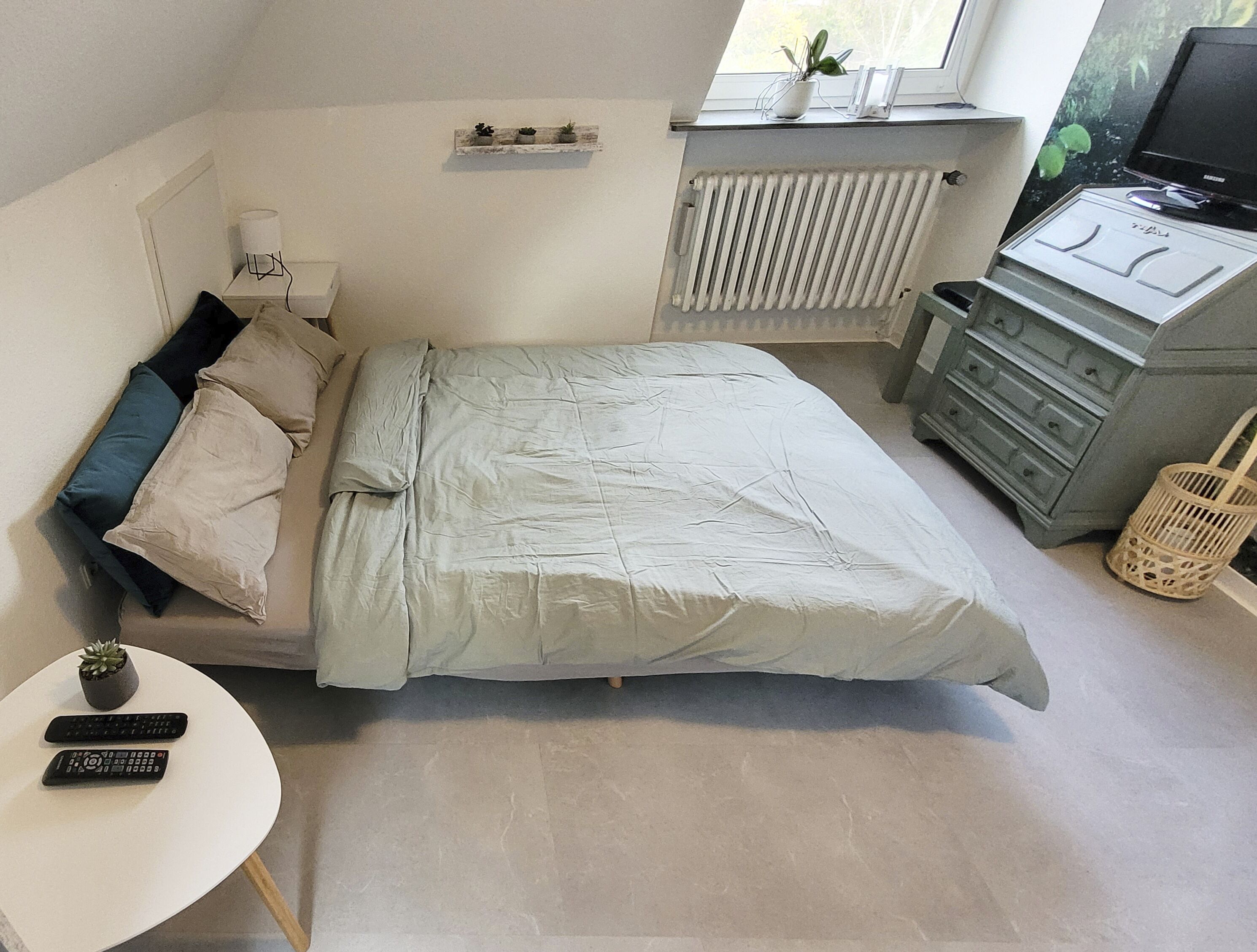 Een slaapkamer met een bed, nachtkastje, kaptafel, radiator en een raam met een plant.