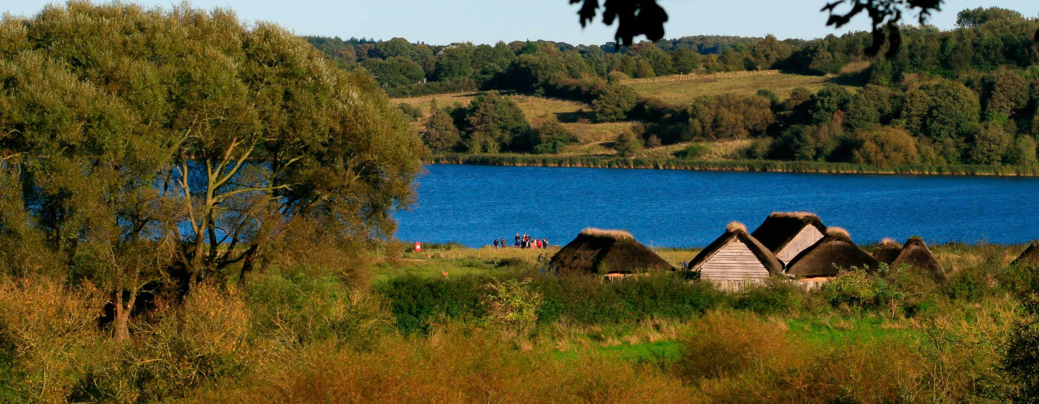 A lakeside view with thatched-roof huts and a group of people by the water.