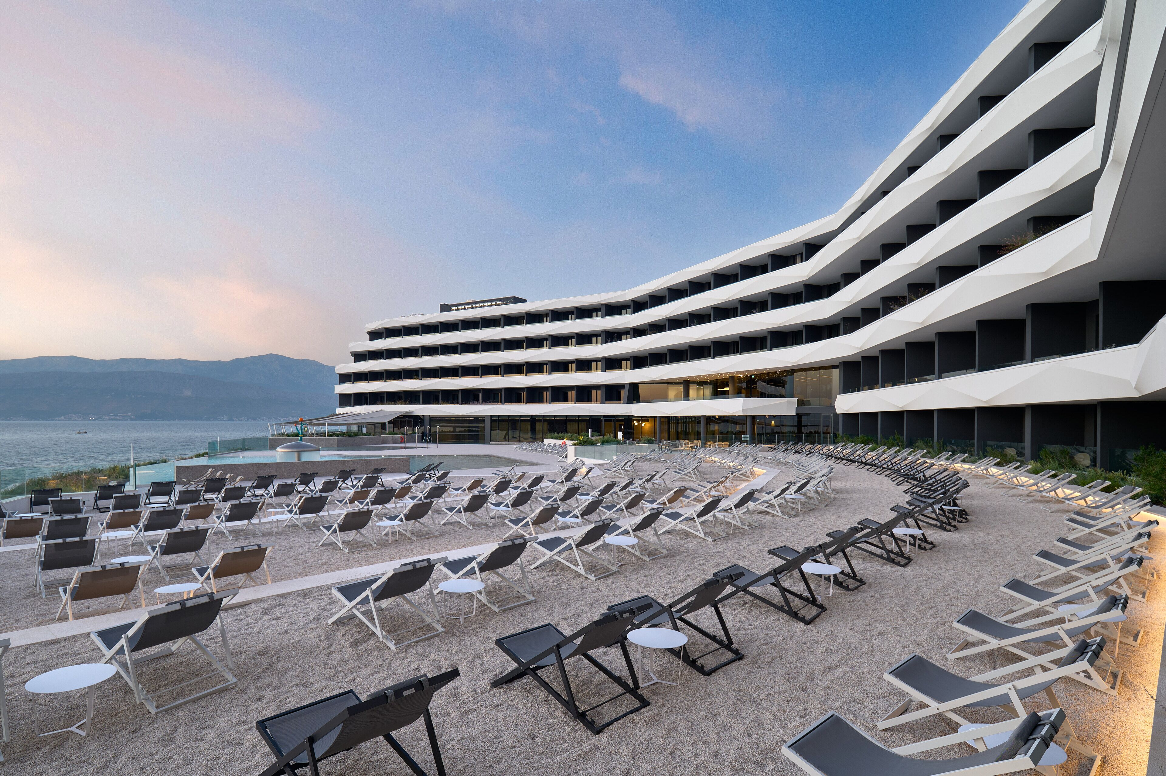 A modern hotel with a curved design, a pool, and sun loungers arranged on a sandy area.
