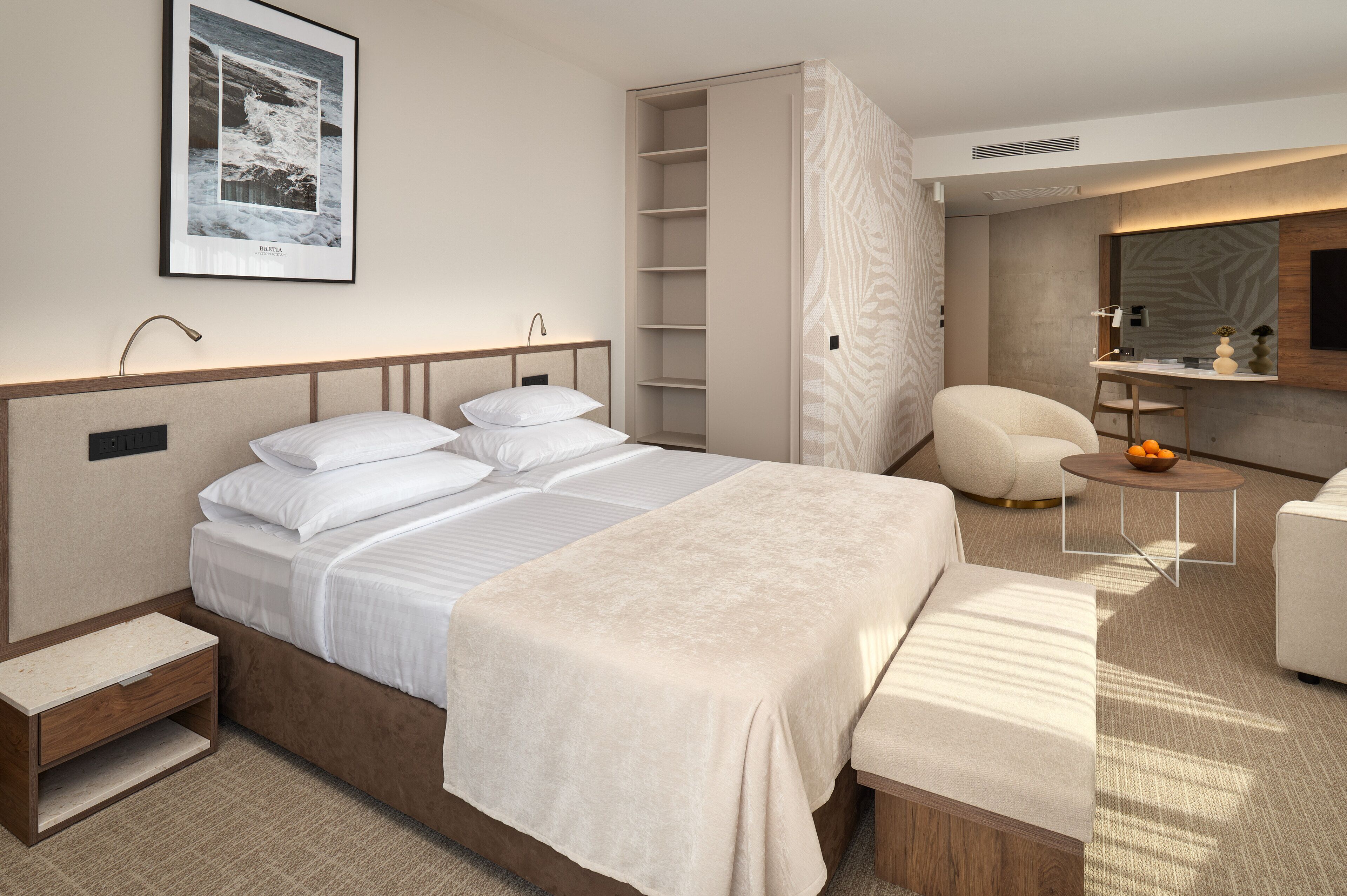 A modern hotel room with a large bed, a seating area, a built-in wardrobe, and a small table with a bowl of fruit.