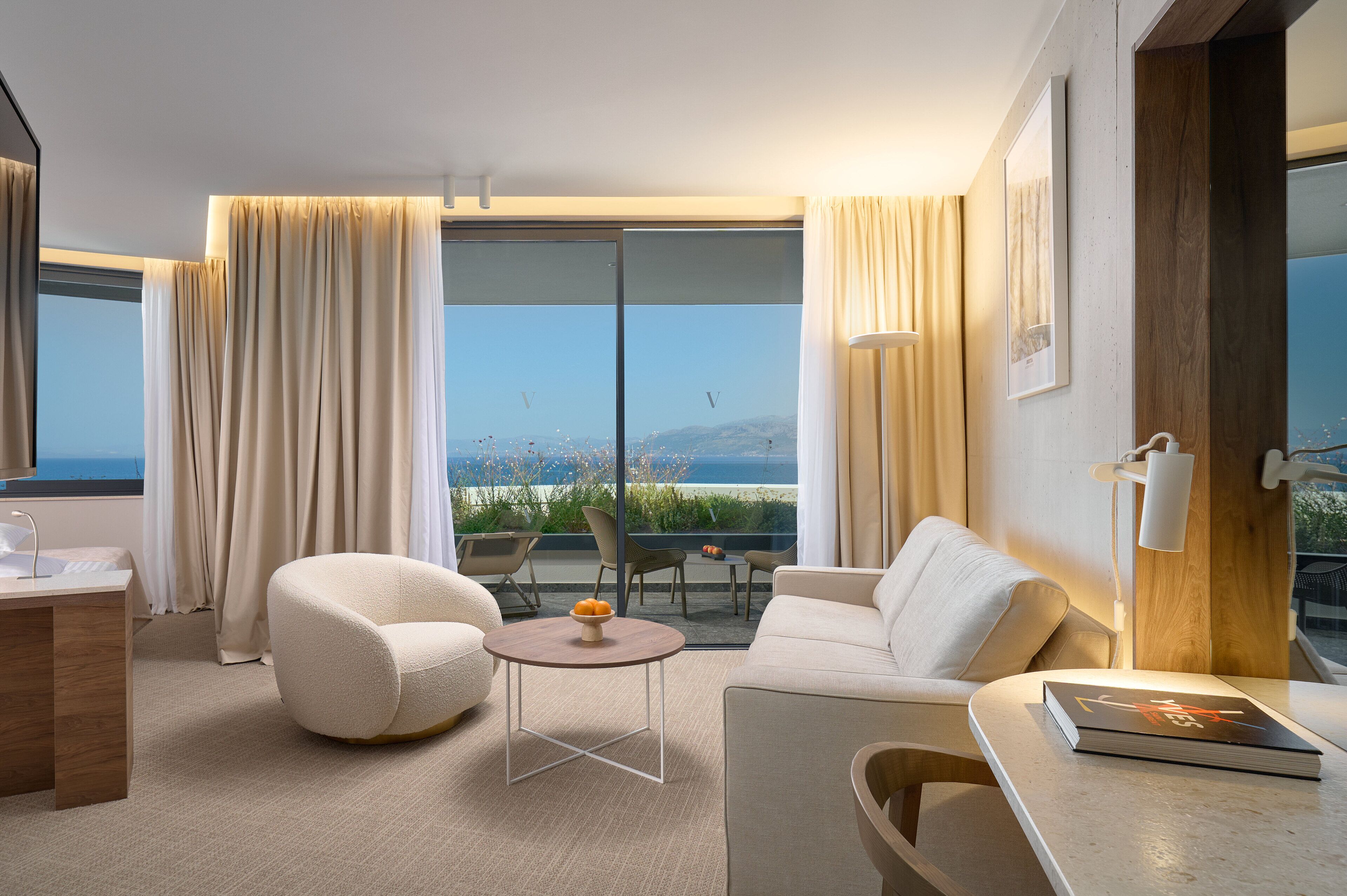 Une chambre d’hôtel moderne avec un canapé, un fauteuil et une petite table sur laquelle se trouve un saladier rempli de fruits. Une grande fenêtre, ornée de rideaux, offre une vue sur la mer, et le sol est en bois.