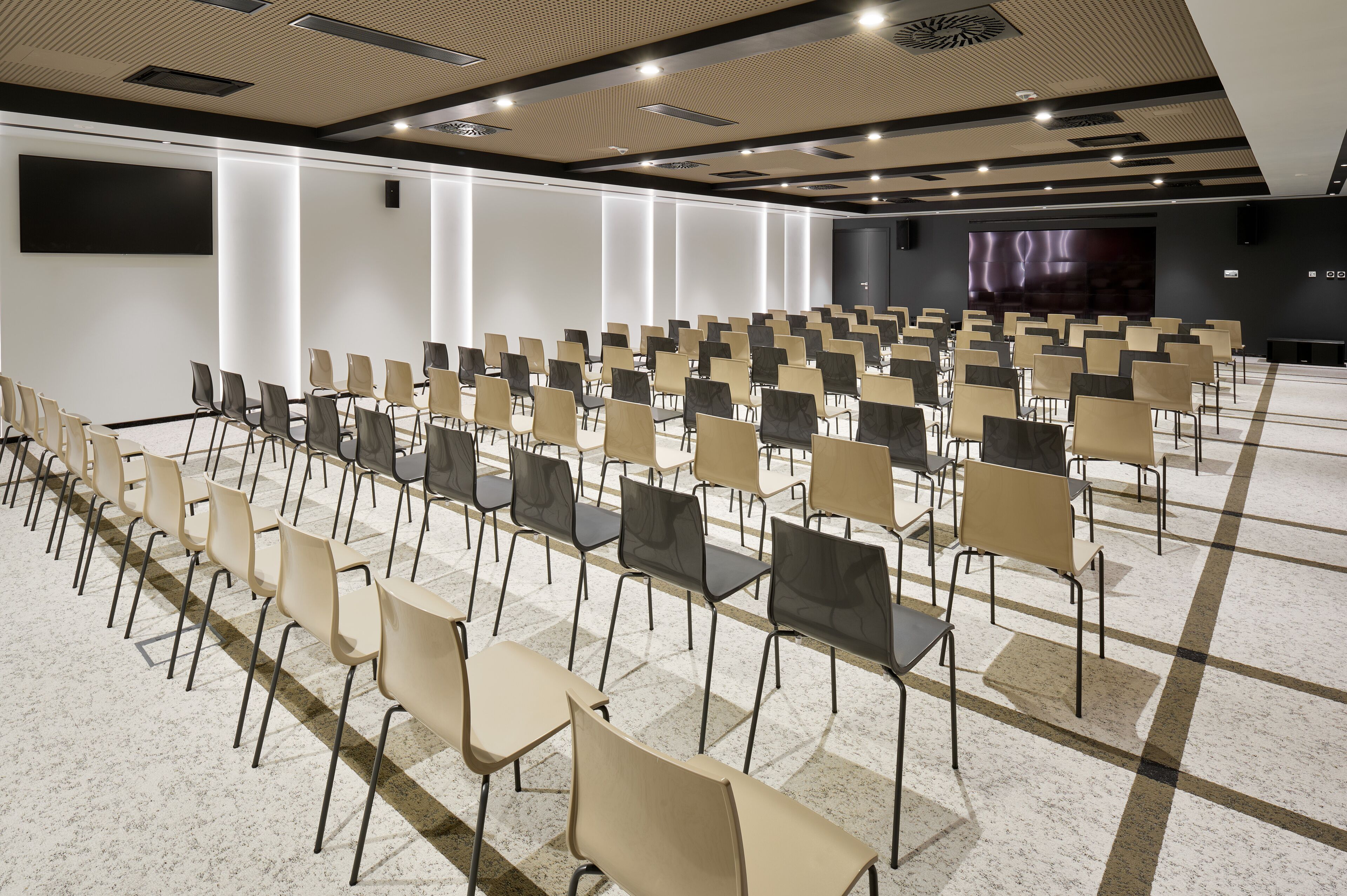 Un salón de conferencias con filas de sillas, una pantalla grande y una pared oscura con un panel iluminado.