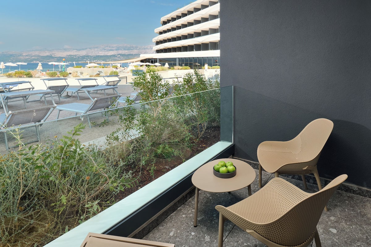 A balcony with wicker chairs, a small table with apples, and a view of a pool and buildings.