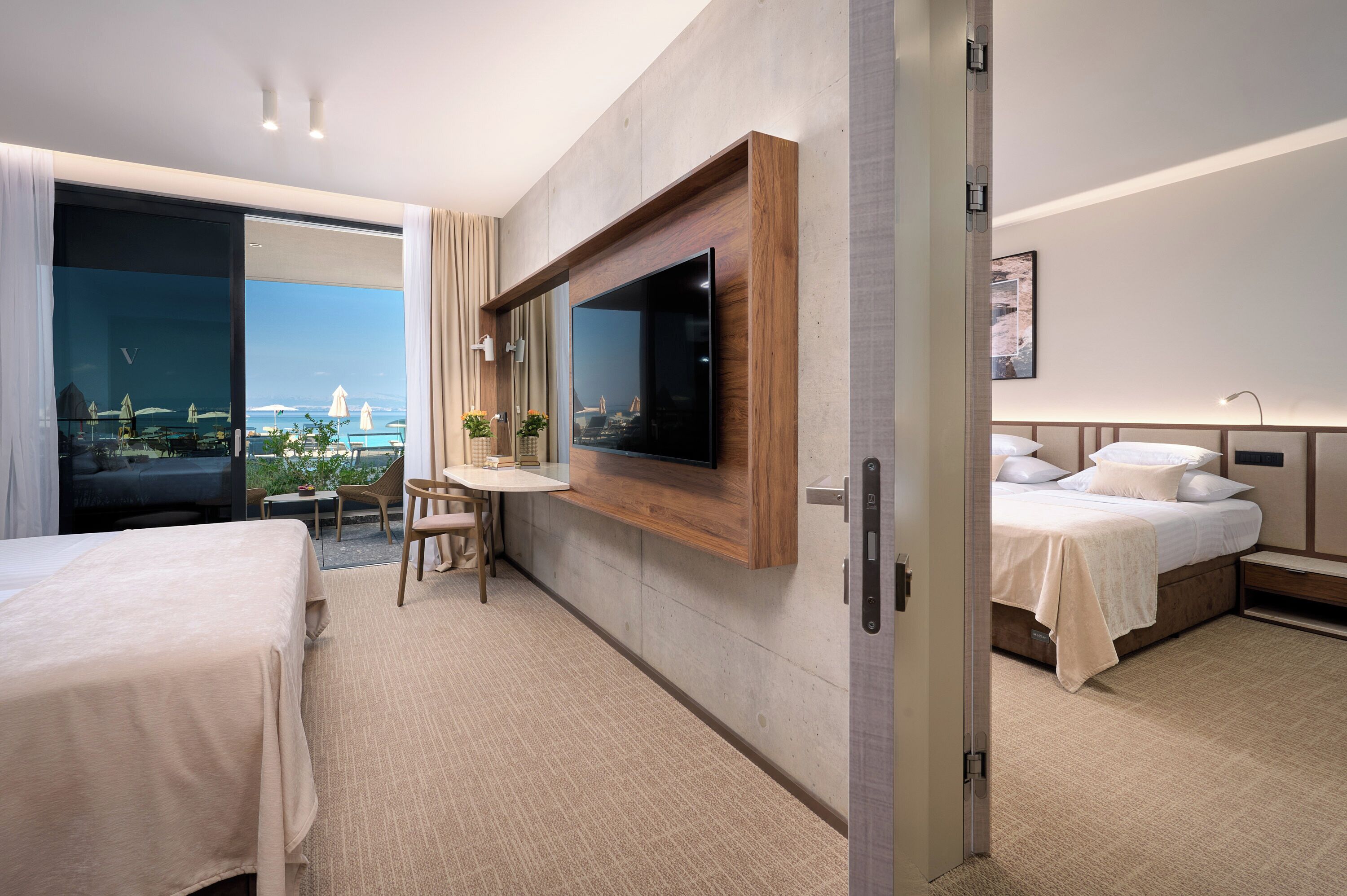 A modern hotel room with a large TV, a bed, a desk, and a balcony with a view of a bridge.