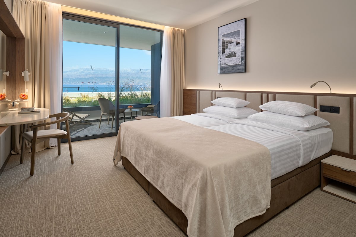 A hotel room with a large bed, a desk with a chair, a small table with fruit, and a view of the sea.