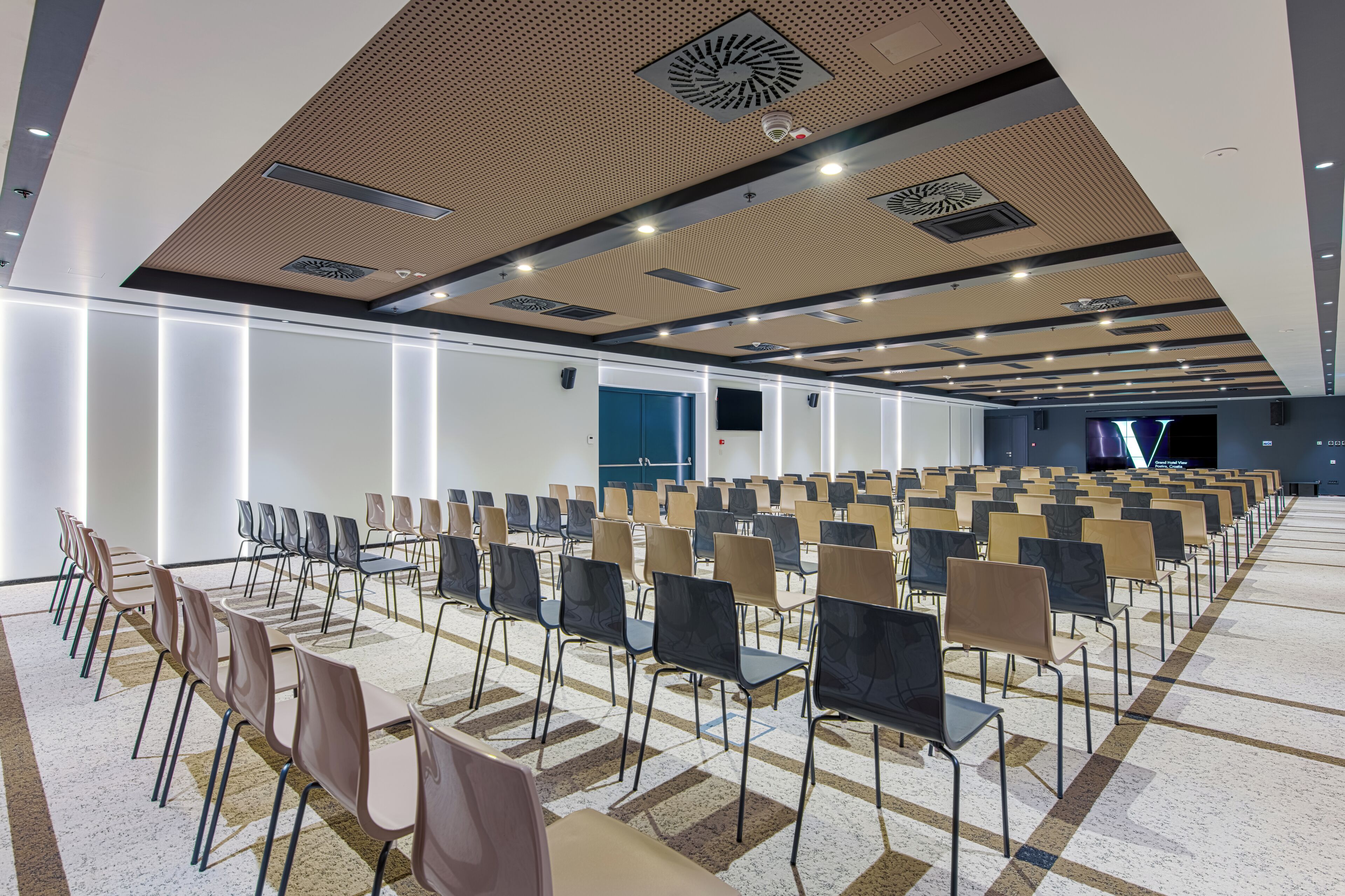 Un salón de conferencias con filas de sillas, un diseño de techo moderno y una pantalla grande al frente.