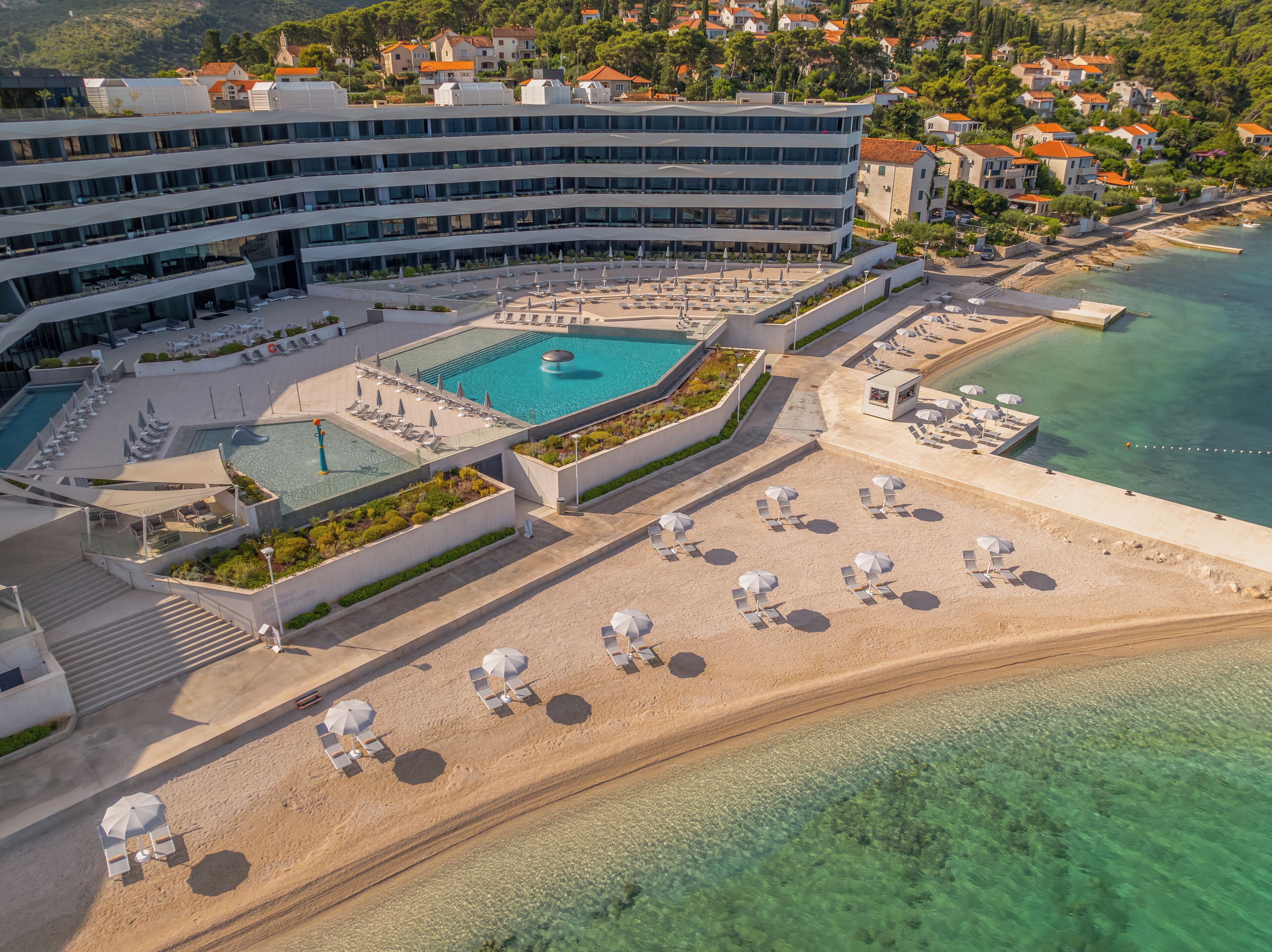 Vista aérea de un hotel con una amplia piscina, zona de playa con tumbonas y una vista despejada del mar.