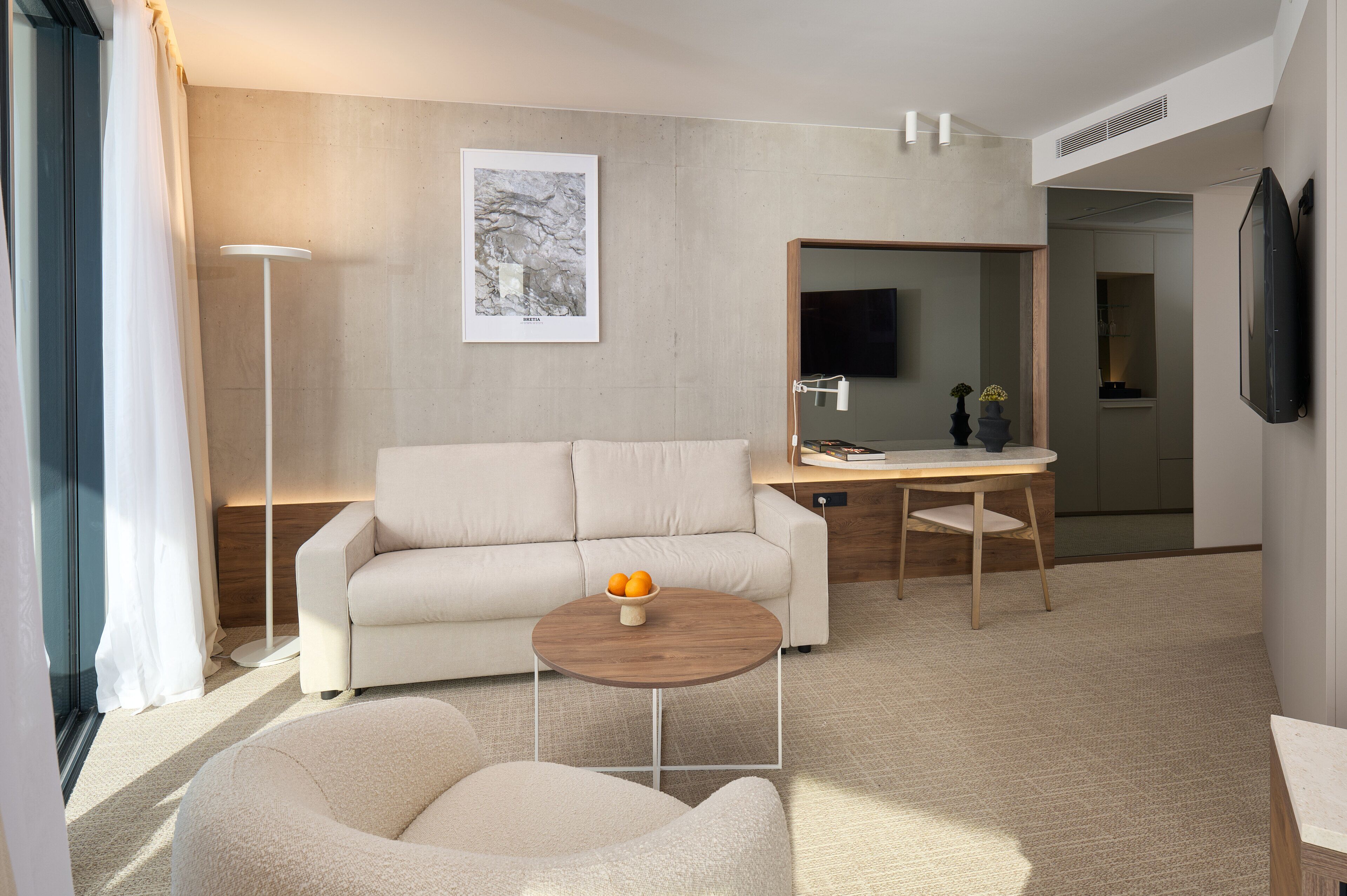 Una habitación de hotel moderna con un sofá, una mesa redonda de madera con un frutero, una lámpara de pie y un espejo grande.