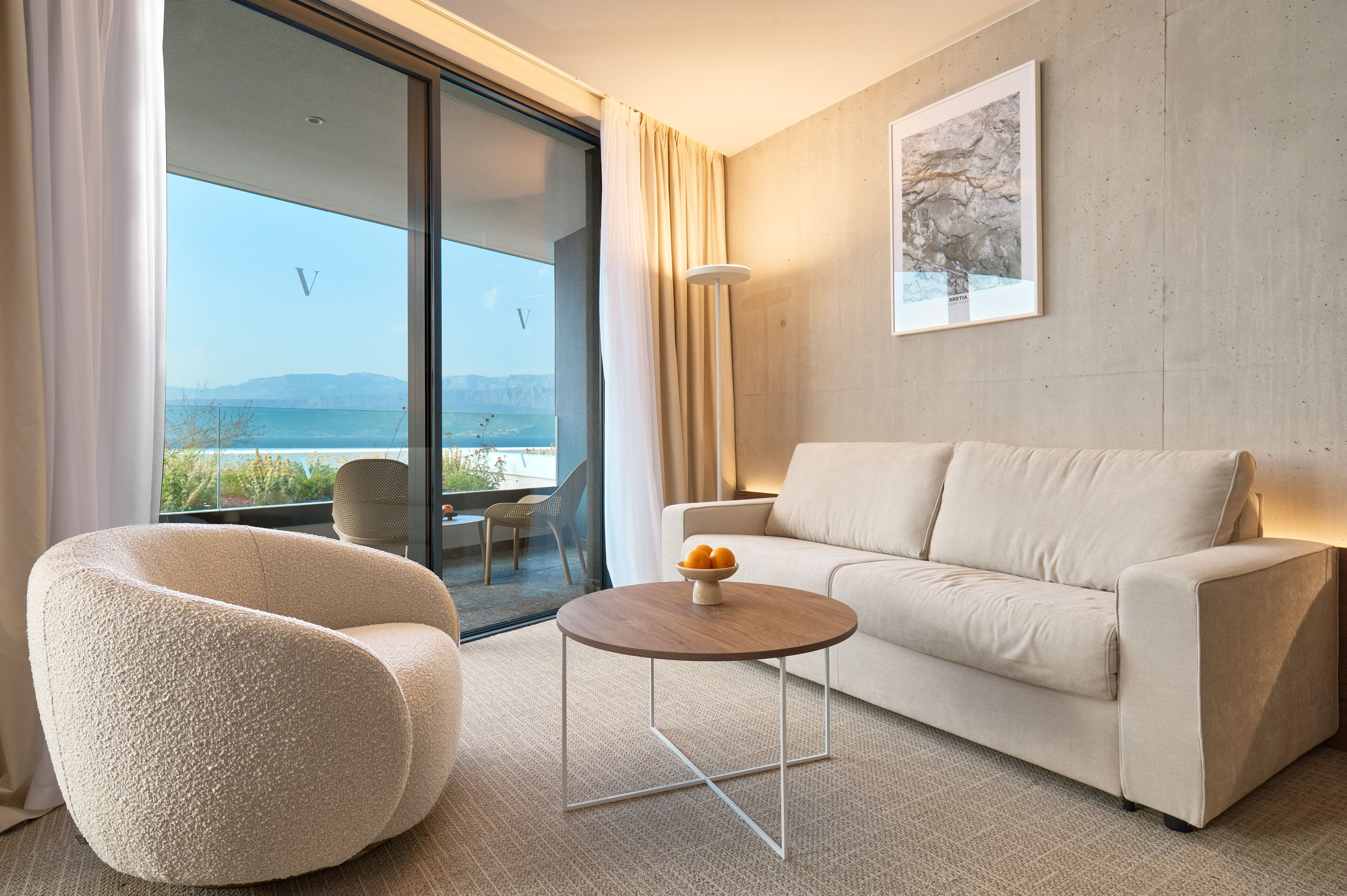 A modern living room with a sofa, armchair, and coffee table, a large window with a view of the sea, and a framed artwork on the wall.