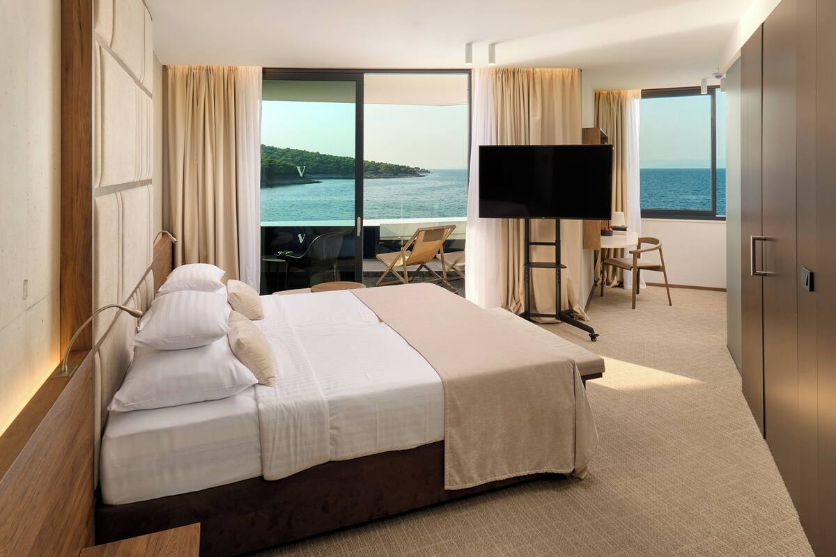 A hotel room with a large bed, a TV, a desk with a chair, and a view of the sea.